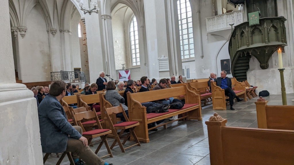 In einer weiß gestrichenen, großen Kirche sitzen Pfarrer in den Kirchenbänken, einer spricht am Mikrofon